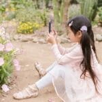 A young girl sits in a garden capturing flowers with her smartphone, surrounded by blossoms.