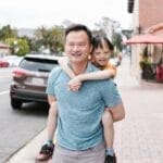 A joyful Asian father giving his child a piggyback ride on a city street.