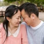 Smiling Asian couple embracing on a sunny day outdoors, sharing a moment of joy.