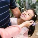 A joyful child smiling as a family member caresses her hair in an outdoor garden.