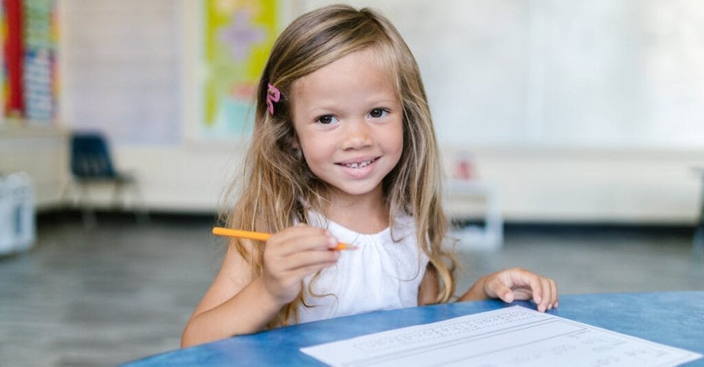 A young girl smiles while working on schoolwork in a bright classroom.