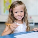 A young girl smiles while working on schoolwork in a bright classroom.