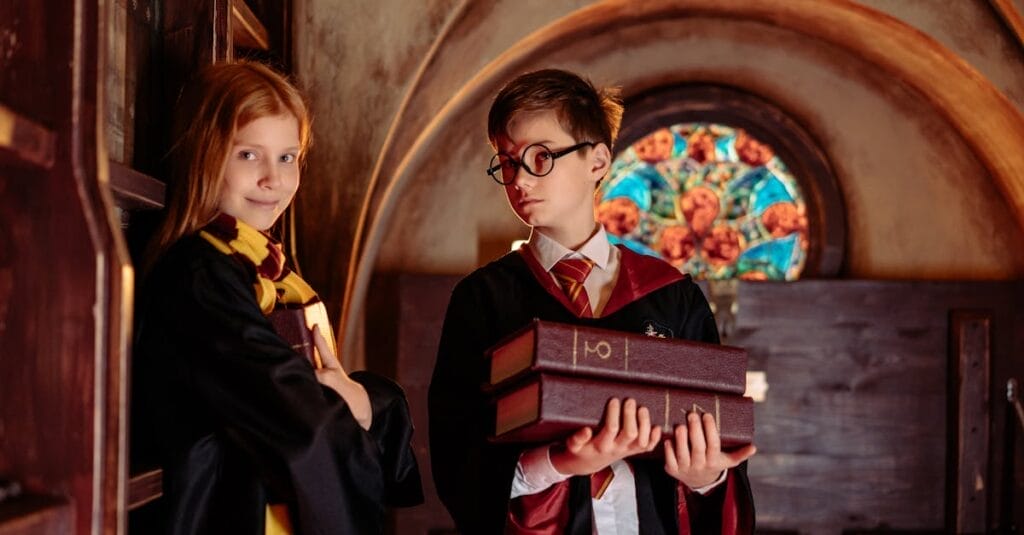 Two children dressed as wizards with robes and glasses, holding books indoors.