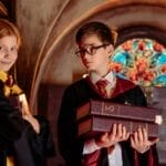 Two children dressed as wizards with robes and glasses, holding books indoors.