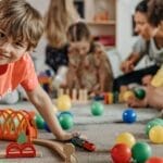 Happy children and a caregiver playing with colorful toys in a lively indoor playroom setting.