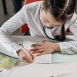 A young girl focused on homework, writing in a notebook in a classroom setting.