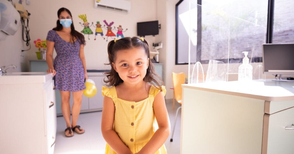 A cheerful child in a yellow dress at a pediatric clinic with doctor in background.