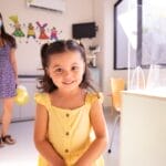 A cheerful child in a yellow dress at a pediatric clinic with doctor in background.