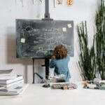 Curious young boy drawing STEM concepts on a blackboard indoors, surrounded by educational materials.