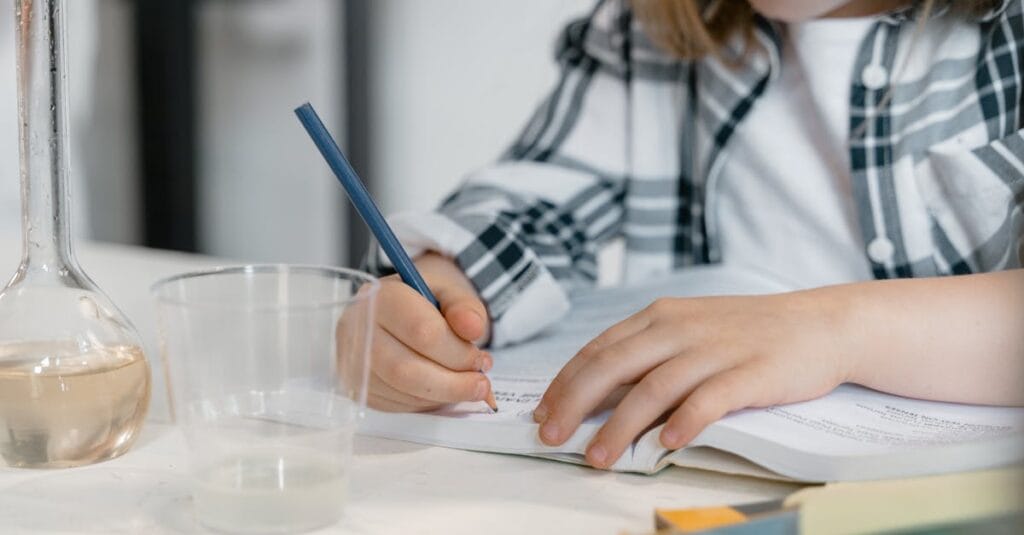 Young child learning science, writing surrounded by books and laboratory beaker.