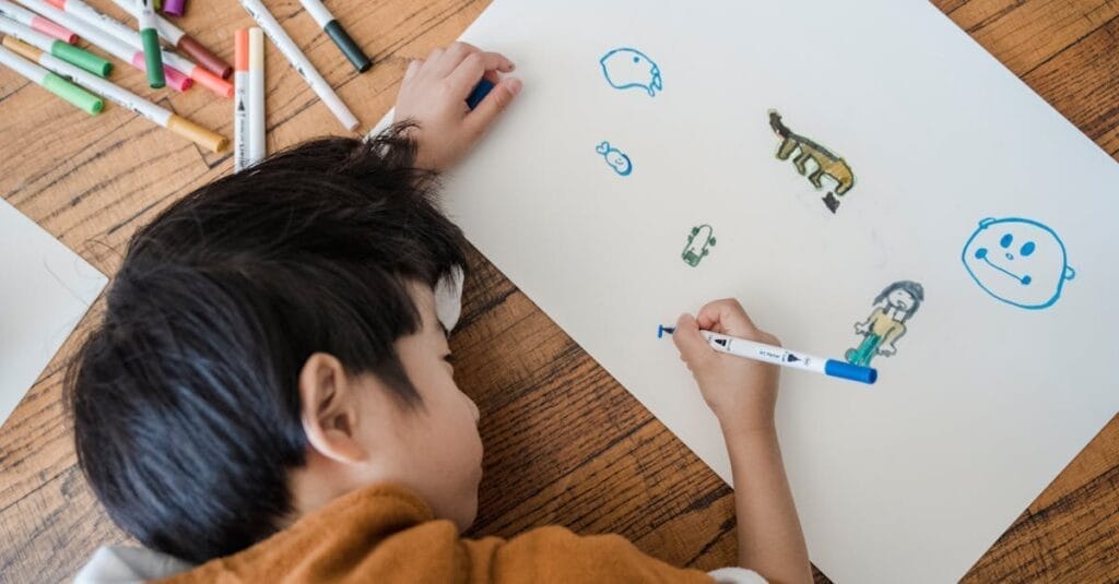 Young child drawing creative images on white paper with colorful pens on a wooden surface.