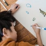 Young child drawing creative images on white paper with colorful pens on a wooden surface.