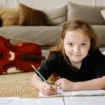 A young girl writing musical notes on the floor next to her cello, creating a cozy home music practice scene.