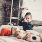 Smiling child with a camera surrounded by stuffed toys in a cozy bedroom setting.