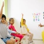 Children raising hands in classroom with a teacher, learning and engaging.