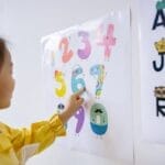 A young girl in a yellow shirt learning numbers in an educational setting, pointing at colorful numbers on a wall poster.