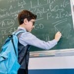 A young boy with glasses solving math equations on a classroom blackboard.