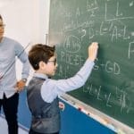 A teacher guides a student solving math equations on a classroom chalkboard.