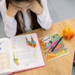 A young student studying at a desk with an open book, colorful notebook, pens, and an apple.