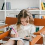 Caucasian girl sitting in a chair drawing with colored pencils in a cozy home library.