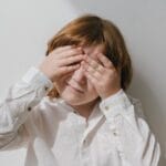 Caucasian child with red hair covers face with hands in bright indoor setting.