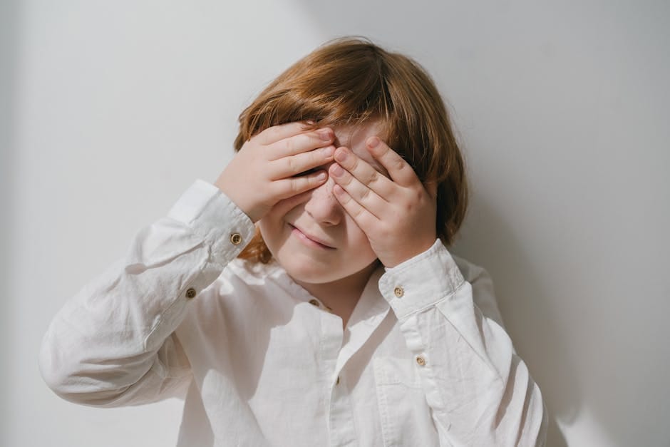 Caucasian child with red hair covers face with hands in bright indoor setting.