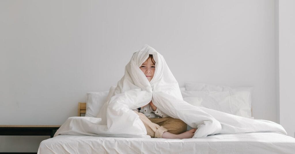 Young child wrapped in a soft white comforter, sitting on a bed in a minimalist room.