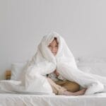 Young child wrapped in a soft white comforter, sitting on a bed in a minimalist room.