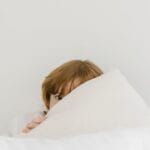 A shy child peeks over a soft white pillow in a cozy indoor setting.