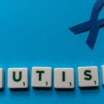 Flat lay of scrabble tiles spelling autism with a blue ribbon on a blue background.