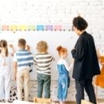 Group of children and teacher interacting with a whiteboard in a bright, indoor classroom setting.