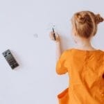 A young girl in an orange dress writes on a whiteboard in a classroom.