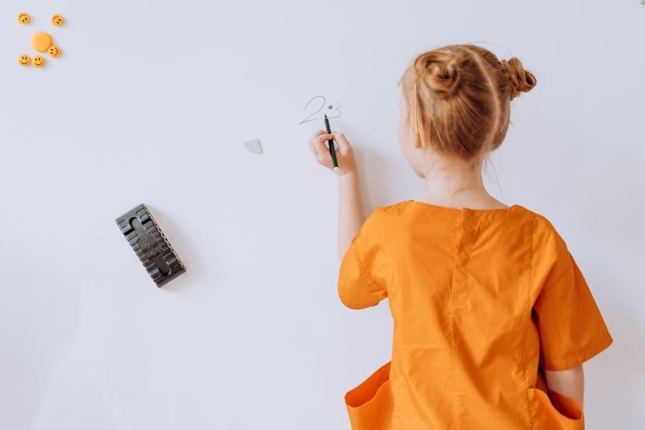 A young girl in an orange dress writes on a whiteboard in a classroom.