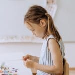 A young girl crafting with colored pencils in a bright, creative classroom setting.