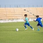 Two boys energetically play soccer on a sunny day in an empty stadium field.