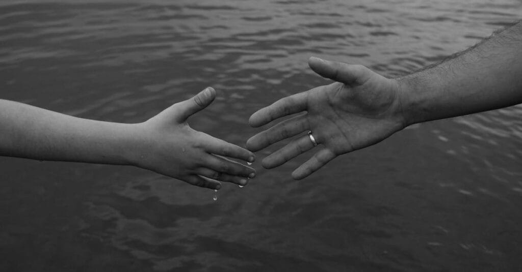 Monochrome image of two hands reaching out over water, symbolizing connection and support.