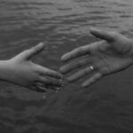 Monochrome image of two hands reaching out over water, symbolizing connection and support.