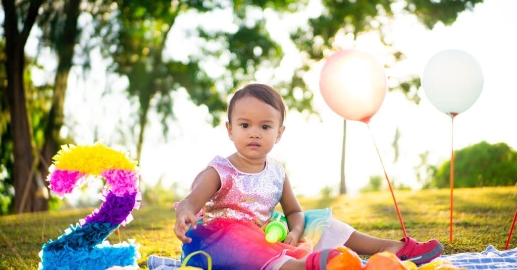 A toddler enjoys a vibrant birthday picnic outdoors with balloons and colorful decorations.