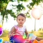 A toddler enjoys a vibrant birthday picnic outdoors with balloons and colorful decorations.