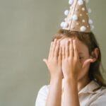 A playful child hides their face with hands while wearing a festive party hat, creating a whimsical moment.