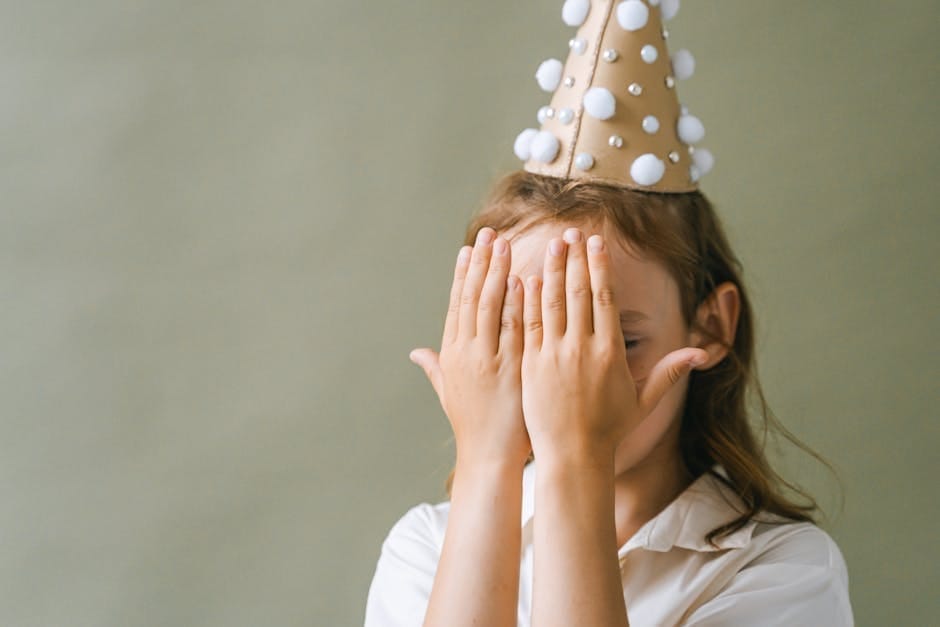 A playful child hides their face with hands while wearing a festive party hat, creating a whimsical moment.