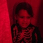 Child in a skeleton costume in a red-illuminated bathroom appears playful and spooky.