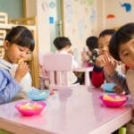 Kids seated around a table in a colorful classroom, eating snacks happily.