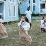 Group of children enjoying a sack race in a backyard setting, surrounded by family.