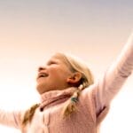 A joyful young girl with braided hair celebrates with arms raised outdoors. Captures happiness and freedom.