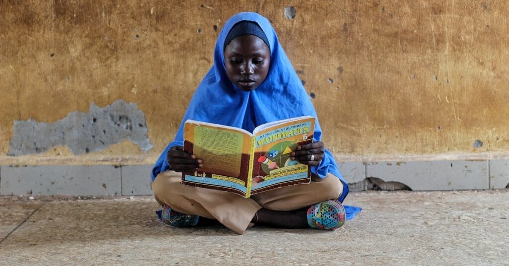 A young girl in hijab sitting on a floor reading a mathematics book intensely.