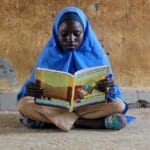 A young girl in hijab sitting on a floor reading a mathematics book intensely.