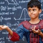 A young boy dressed as a superhero stands confidently in front of a chalkboard filled with math equations.