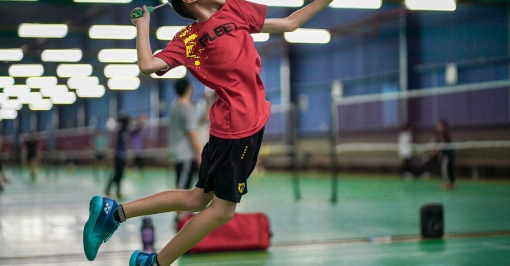 A young boy enthusiastically jumps to hit a badminton shuttlecock inside a sports hall.