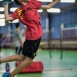 A young boy enthusiastically jumps to hit a badminton shuttlecock inside a sports hall.